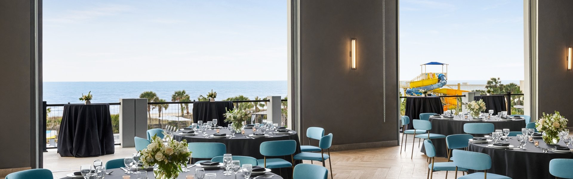 Seaside Vista ballroom above Black Drum Brewing set up in rounds for a banquet event. The garage doors are open with a direct view of the ocean and the terrace has cocktail tables set up.