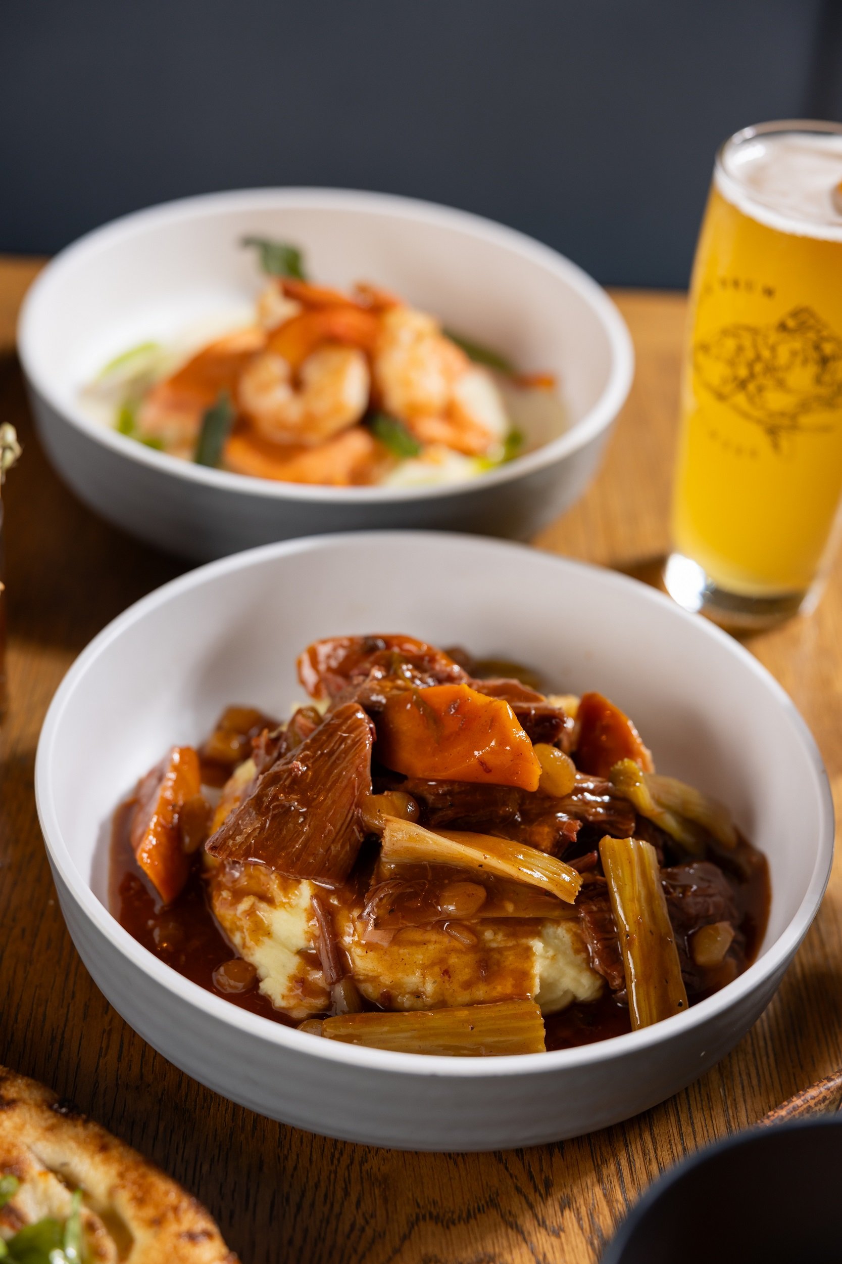 2 bowls of food and a beer on the table. One bowl has Pot Roast and the other has ravioli with shrimp