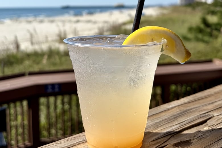 Plastic cup with ice and a lemon slice on a weathered wooden railing at a sunny beach, ocean and buildings in the background.