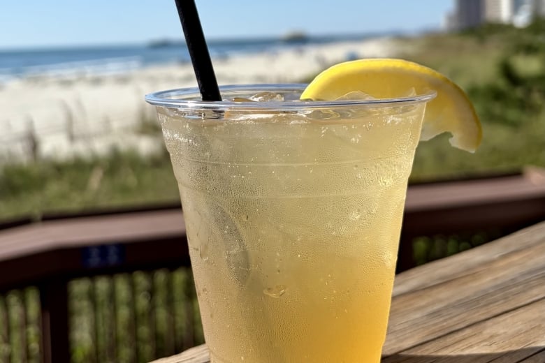 Clear plastic cup of lemonade with ice and a lemon wedge, on a weathered wooden table by the beach under a bright blue sky.
