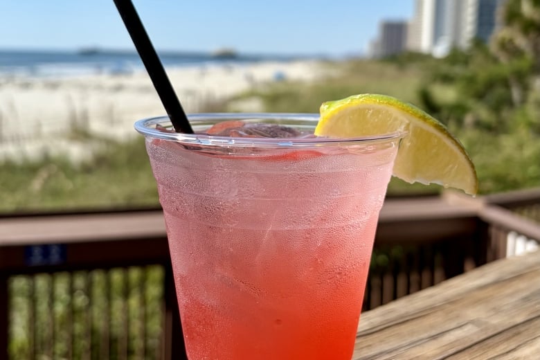 Pink iced drink in a clear cup with a black straw and lime wedge on a wooden railing, with a beach and high-rise buildings in the background.