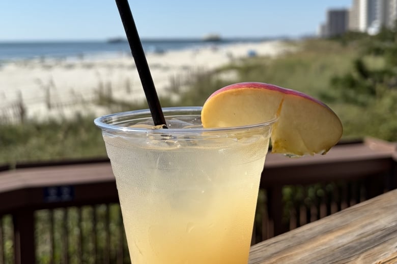 A pale yellow cocktail in a clear plastic cup with a black straw and a lemon wedge on the rim, set on a wooden railing with a sunny beach and blue sky in the background.
