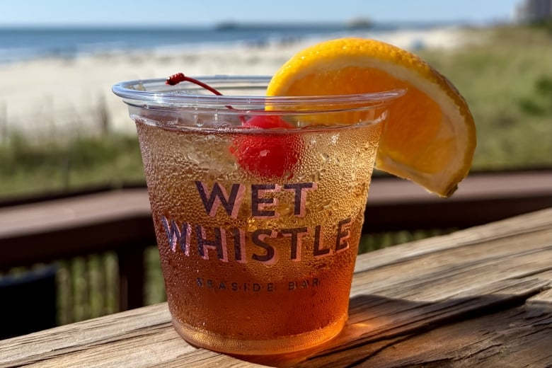 Ice-cold drink in a plastic cup with an orange wedge, on a wooden table at the beach under a clear blue sky.