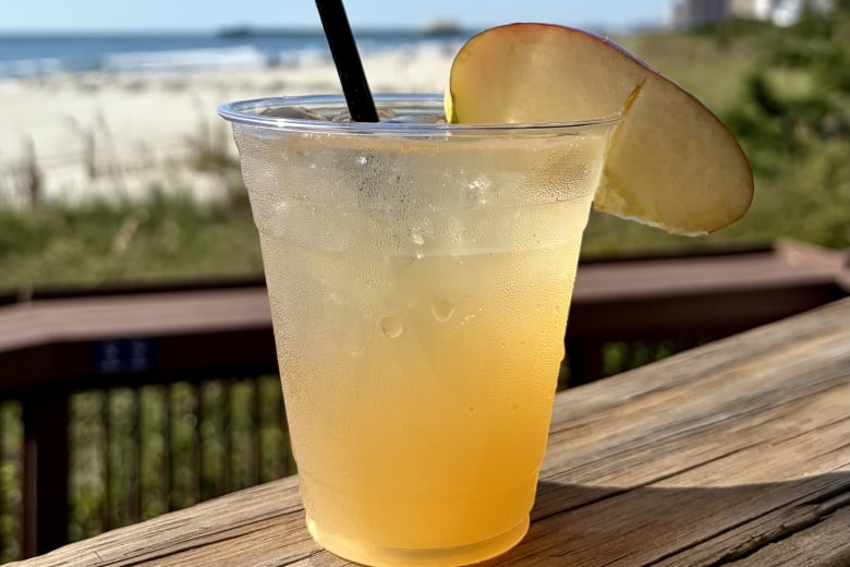 Cold lemonade in a clear plastic cup with a black straw and lemon wedge on a wooden table by the beach. Readable and refreshing scene at the coast.